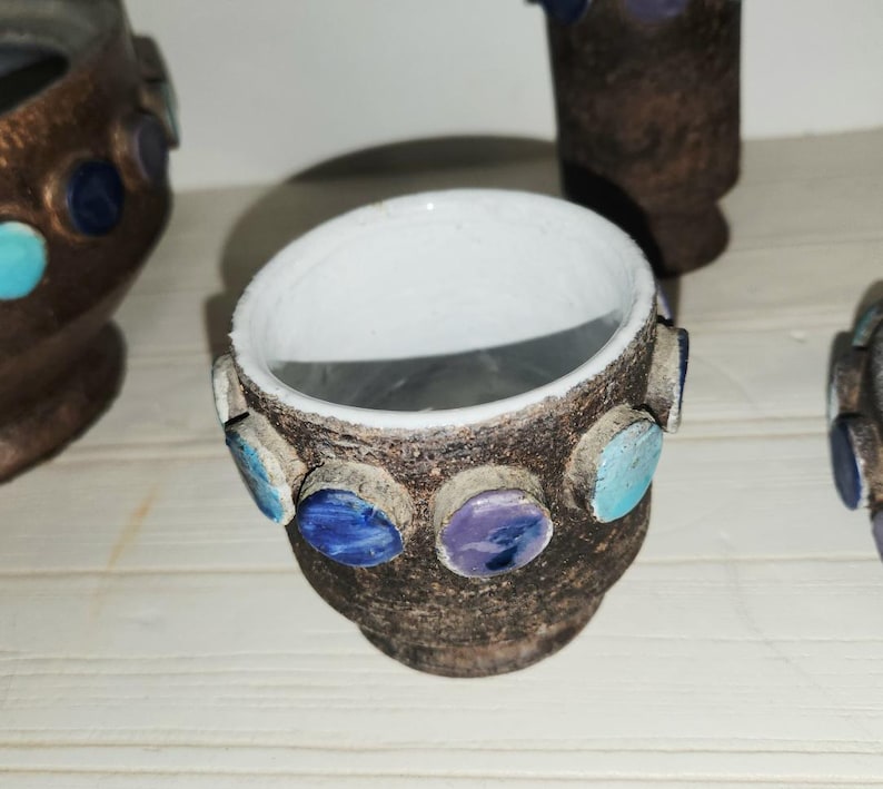 May include: A small, handcrafted ceramic pot with a dark brown exterior and a white interior. The pot is adorned with colorful, circular accents in shades of blue, turquoise, and purple. The pot is likely used for decorative purposes.