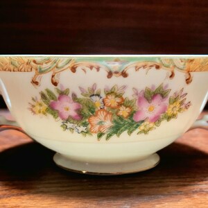 May include: A white porcelain soup bowl with two handles. The bowl has a floral design with pink, yellow, and orange flowers on a green background. The rim of the bowl is decorated with gold trim.