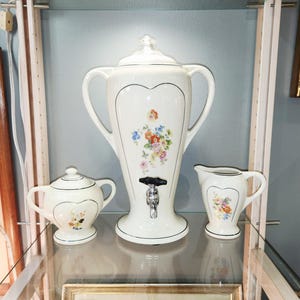 Peut inclure: Un distributeur de boissons en céramique crème avec un couvercle en verre, deux anses et un robinet noir et argent, accompagné d'un sucrier assorti avec couvercle et d'un pot à lait, ornés de délicats motifs floraux multicolores.