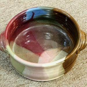 May include: A round ceramic baking dish with two handles. The dish is glazed in shades of brown, green, pink, and blue.