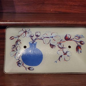 May include: Rectangular light switch cover with a floral design. A blue vase holds branches with white and red flowers. The cover has two holes for the switch and a black border. The background is a light cream color.