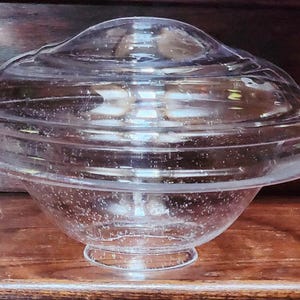 May include: A clear glass bowl with a matching lid. The bowl has a rounded base and a flared rim. The lid is domed and fits snugly on top. The glass has a slightly bubbly texture, giving it a vintage look.