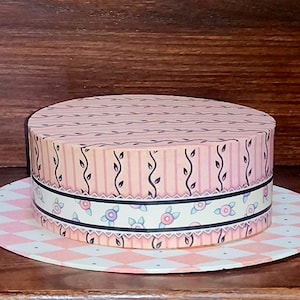 Peut inclure: Une boîte ronde décorative avec un couvercle, ornée d'un motif floral et géométrique rose et blanc. La boîte a une base assortie, et le design comprend des rayures, des vignes et des motifs floraux. La boîte est sur une surface en bois.