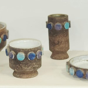 May include: A collection of brown ceramic pottery pieces, including a lidded jar, a small cup, a vase, a candle holder, and a small dish. Each piece is adorned with a row of blue and turquoise circular accents. The pottery has a textured surface.