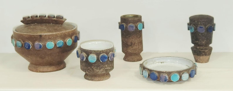 May include: A collection of brown ceramic pottery pieces, including a lidded jar, a small cup, a vase, a candle holder, and a small dish. Each piece is adorned with a row of blue and turquoise circular accents. The pottery has a textured surface.