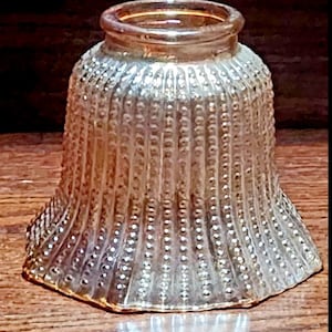 May include: A clear glass lampshade with a textured, ridged design. The shade has a flared shape with a rounded top and a series of vertical ridges and small, raised dots. The glass has a slightly amber hue, and the shade sits on a wooden surface.