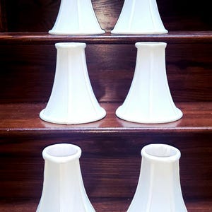 Op de afbeelding: Zes witte, klokvormige lampenkappen zijn gerangschikt op houten planken. De kappen hebben een glad, licht getextureerd oppervlak en een klassiek ontwerp. De kappen zijn een neutrale kleur, geschikt voor diverse interieurstijlen.