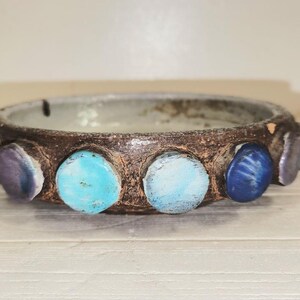 May include: A handcrafted ceramic bowl with a dark brown exterior and a light gray interior. The rim is adorned with colorful, round stones in shades of blue, turquoise, and purple. The bowl is a decorative piece.