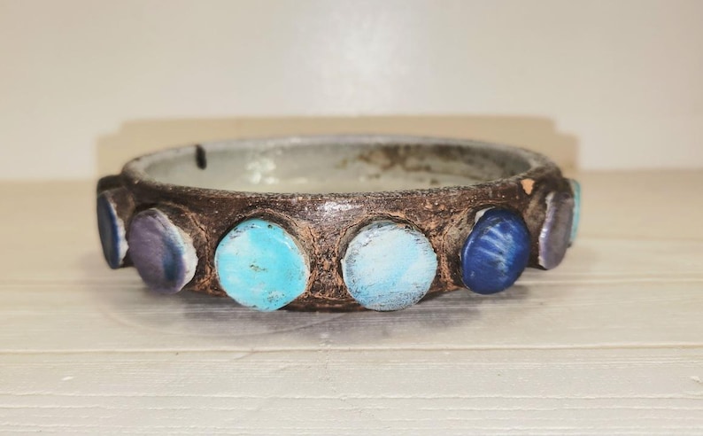 May include: A handcrafted ceramic bowl with a dark brown exterior and a light gray interior. The rim is adorned with colorful, round stones in shades of blue, turquoise, and purple. The bowl is a decorative piece.