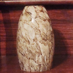 May include: An egg-shaped decorative vase with a beige and brown feather-like pattern. The vase has a glossy finish and is set against a dark brown wooden background. The vase is a decorative item.