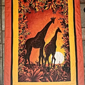 May include: A quilted wall hanging with a yellow and orange border features a silhouette of two giraffes standing in a field of brown and green foliage against a sunset background.