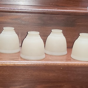 May include: A set of four bell-shaped lamp shades made of frosted white glass. Each shade features a smooth, matte finish with a slightly flared bottom edge and a narrow opening at the top for light fixture attachment, arranged in a row.