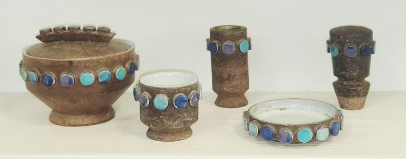 May include: A collection of brown ceramic pottery pieces, including a lidded jar, a small cup, a vase, a candle holder, and a small dish. Each piece is adorned with a row of blue and turquoise circular accents. The pottery has a textured surface.