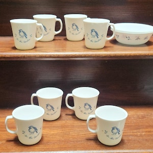 May include: A collection of off-white mugs and a bowl with a blue bird design. The mugs have a curved handle and a bird illustration with the text "In the Garden". The bowl has a lid and a similar bird design.
