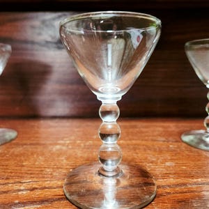 May include: Clear glass cordial glasses with a round base and a stem made of stacked spheres. The bowl of the glass is conical. The glasses are on a wooden surface.