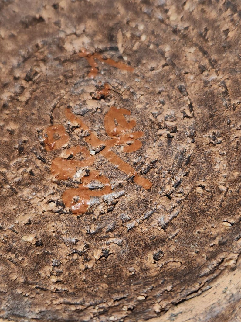 May include: Close-up of a textured, brown ceramic surface with a circular pattern. The surface has a rough, uneven texture with small, dark spots. There is a reddish-brown, raised design in the center, resembling characters.