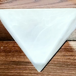Puede incluir: Un jabonera triangular blanca con apariencia marmolada. La jabonera está hecha de un material sólido y tiene una superficie lisa. La jabonera está diseñada para sostener una pastilla de jabón y se coloca sobre una superficie de madera.
