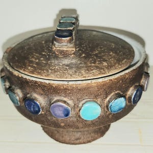 May include: A brown ceramic lidded bowl with a handle. The bowl is decorated with colorful, circular accents in shades of blue, teal, and purple. The lid and base have a textured appearance.