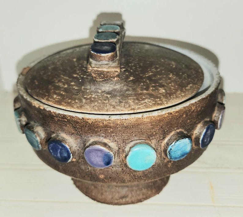May include: A brown ceramic lidded bowl with a handle. The bowl is decorated with colorful, circular accents in shades of blue, teal, and purple. The lid and base have a textured appearance.