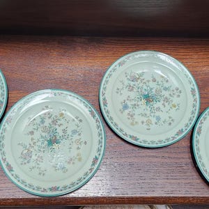 Op de afbeelding: Vier ronde, lichtgroene borden met een bloemmotief. Elk bord heeft een turquoise rand en een centraal bloemmotief in tinten blauw, geel en roze. De borden zijn gerangschikt op een donkerbruin houten oppervlak.