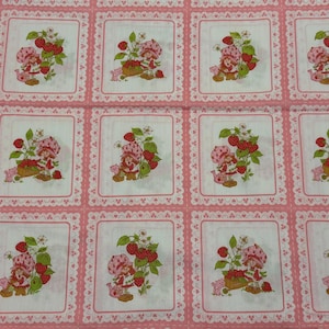 May include: Pink and white fabric featuring a repeating pattern of a cartoon character with strawberries. Each square has a pink border with a white lace design. The character is wearing a pink hat and dress, holding a basket of strawberries.
