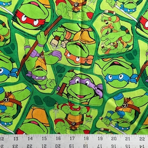 May include: Green fabric featuring a repeating pattern of the Teenage Mutant Ninja Turtles. Each turtle is depicted in a dynamic pose, wearing their signature colored masks and holding weapons. The background is a lighter green with a grid-like design.