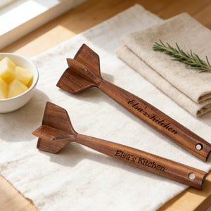 May include: Two wooden herb choppers with a distinctive design, featuring a blade-like head and a handle engraved with "Elsa's Kitchen." The choppers are made of dark wood and rest on a white cloth, with a bowl of diced potatoes nearby.