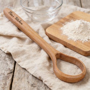 May include: A wooden spoon with a looped handle, engraved with "Elsa's Kitchen." The spoon is on a beige cloth, near a small wooden chopping board with flour and a clear glass bowl. The spoon is made of light brown wood.