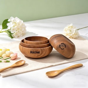 May include: A set of three wooden bowls, one with the name "Rachel" engraved, and a wooden lid. Two wooden spoons are also present. The bowls and spoons are made of a light brown wood. White flowers and a white cloth are in the background.