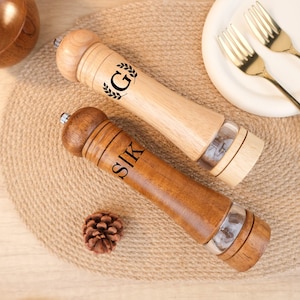 May include: Two wooden salt and pepper grinders. One is light-colored with the letter "G" and a laurel wreath, the other is dark-colored with the letters "SIK". Both have a clear section to view contents.