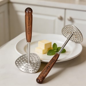 May include: Two stainless steel potato mashers with wooden handles. The handles are engraved with "Elsa's Kitchen". One masher has a round head, the other has a triangular head. The mashers are on a white plate with butter and mint.