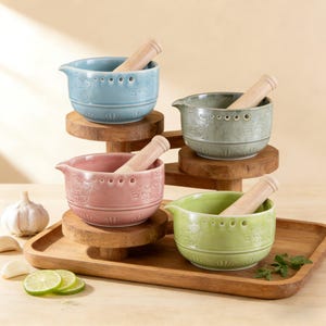 Peut inclure: Quatre ensembles de mortiers et pilons en céramique aux couleurs pastel : bleu, vert, rose et vert clair. Chaque bol a un bec verseur et est associé à un pilon en bois. Les bols sont sur des supports et un plateau en bois.