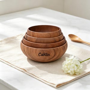 May include: Four wooden bowls of different sizes, nested together. The largest bowl is engraved with the name "Caitlin". A wooden spoon and a white flower are on a beige linen cloth. The bowls are made of wood.