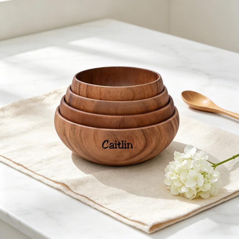 May include: Four wooden bowls of different sizes, nested together. The largest bowl is engraved with the name "Caitlin". A wooden spoon and a white flower are on a beige linen cloth. The bowls are made of wood.