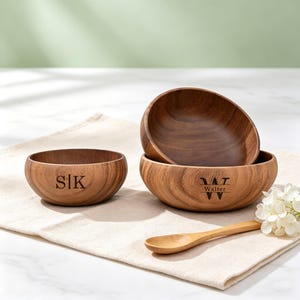 Könnte beinhalten: Drei Holzschalen mit natürlichem Finish werden auf einem beigen Tuch präsentiert. Eine Schale hat die Initialen "SIK" eingraviert, während eine andere "W T Walter" eingraviert hat. Ein kleiner Holzlöffel und weiße Blumen sind ebenfalls vorhanden.