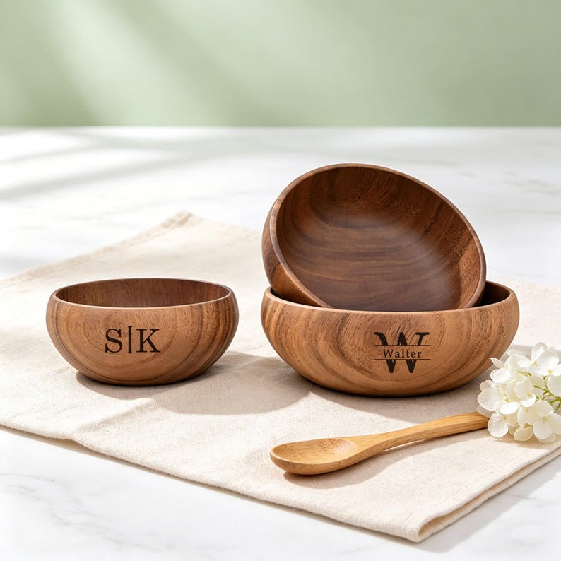May include: Three wooden bowls with a natural finish are displayed on a beige cloth. One bowl has the initials "SIK" engraved, while another has "W T Walter" engraved. A small wooden spoon and white flowers are also present.