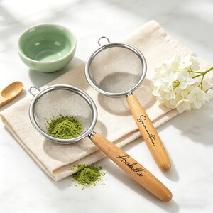 May include: Two stainless steel tea strainers with wooden handles, one filled with green powder. The handles are engraved with the names "Arabella" and "Samantha". A small green bowl and white flowers are also in the image.