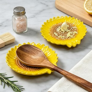 May include: A pair of bright yellow sunflower-shaped kitchen utensils. One holds a wooden spoon, another has grated garlic. A glass salt shaker and a small brush are also visible. The items are on a marble surface.