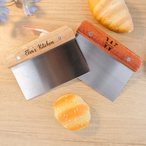 May include: Two stainless steel dough scrapers with wooden handles. One scraper has "Elsa's Kitchen" engraved on the handle, and the other has "Walter" engraved. A small bread roll and a croissant are also visible.