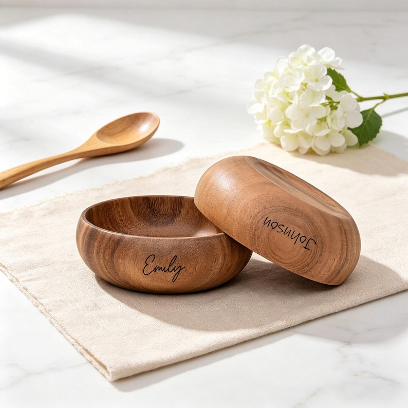 May include: Two wooden bowls, one with "Emily" and the other with "Johnson" engraved. The bowls are light brown and rest on a beige cloth. A wooden spoon and white flowers are also visible in the image.