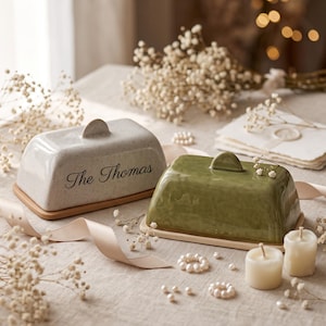 Könnte beinhalten: Zwei rechteckige Butterdosen mit Deckeln und Holzböden. Eine Dose ist gesprenkelt cremeweiß mit dem Text The Thomas in schwarzer Schrift. Die andere Dose ist einfarbig olivgrün. Beide haben einen kleinen halbkreisförmigen Griff am Deckel.