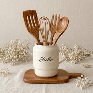 May include: A cream ceramic utensil pot with the name "Stella" in black script, containing wooden kitchen utensils. The utensils include a slotted spatula, whisk, fork, and spoon. The pot rests on a wooden chopping board.