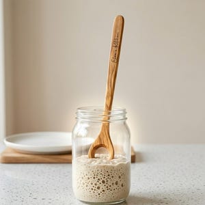 May include: A wooden spoon with the text "Elsa's Kitchen" is placed in a glass jar filled with a bubbly, light brown mixture. The spoon has a unique cutout design. A white plate and wooden cutting board are in the background.