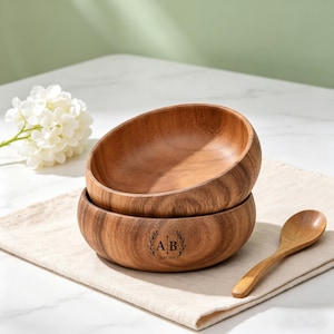 May include: Two wooden bowls stacked on a beige linen napkin, with a wooden spoon. The bottom bowl has a wreath with the initials "AB" and the date "23.07.2025" engraved. A white flower and a white marble surface are in the background.