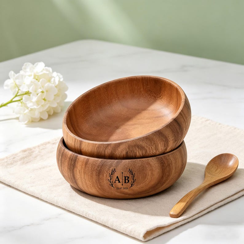 May include: Two wooden bowls stacked on a beige linen napkin, with a wooden spoon. The bottom bowl has a wreath with the initials "AB" and the date "23.07.2025" engraved. A white flower and a white marble surface are in the background.