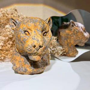 May include: A sculpted bust of a big cat head featuring a mottled texture with a blend of earthy orange and dark grey tones. The feline figure displays detailed facial features including pointed ears, almond-shaped eyes, and a textured snout.