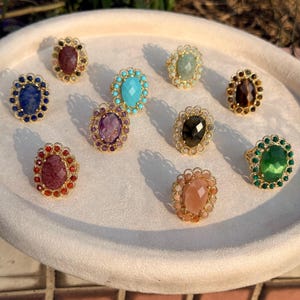 May include: A collection of nine oval-shaped rings featuring faceted gemstones in various colors including blue, purple, red, turquoise, green, brown, and peach. Each ring has a gold-toned metal setting with a decorative beaded edge border.