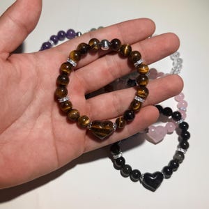 May include: A stretch bracelet featuring brown tiger eye beads, a central tiger eye heart-shaped bead, silver-toned spacers, and small clear crystal accents. The bracelet is arranged in a circular shape with a smooth, polished finish throughout.