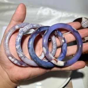 Peut inclure: Quatre bracelets joncs circulaires en pierre polie. Les bracelets présentent des nuances variées de bleu, de lavande et de blanc avec des motifs distincts circulaires, nuageux et marbrés sur les surfaces minérales lisses et translucides.
