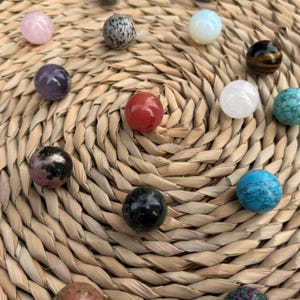 Peut inclure: Une collection de douze pierres précieuses sphériques et lisses de couleurs et textures variées, incluant rose clair, gris tacheté, blanc, brun foncé, violet, rouge, transparent, turquoise, noir et bleu, disposées en cercle.